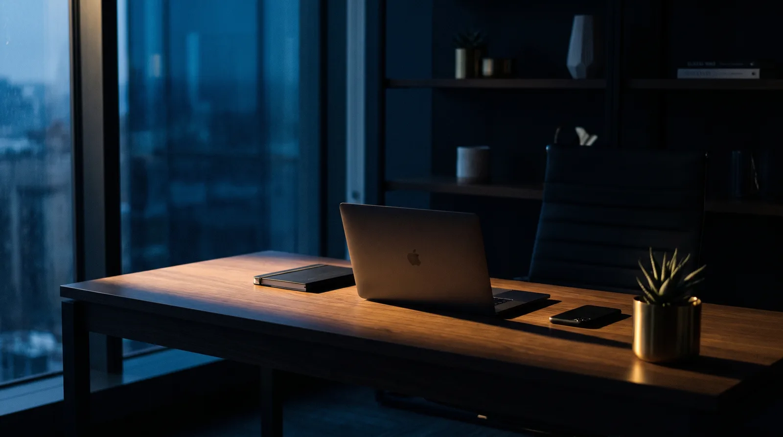 Clean, orderly desk by tall windows at dusk — laptop, notebook, and phone in place